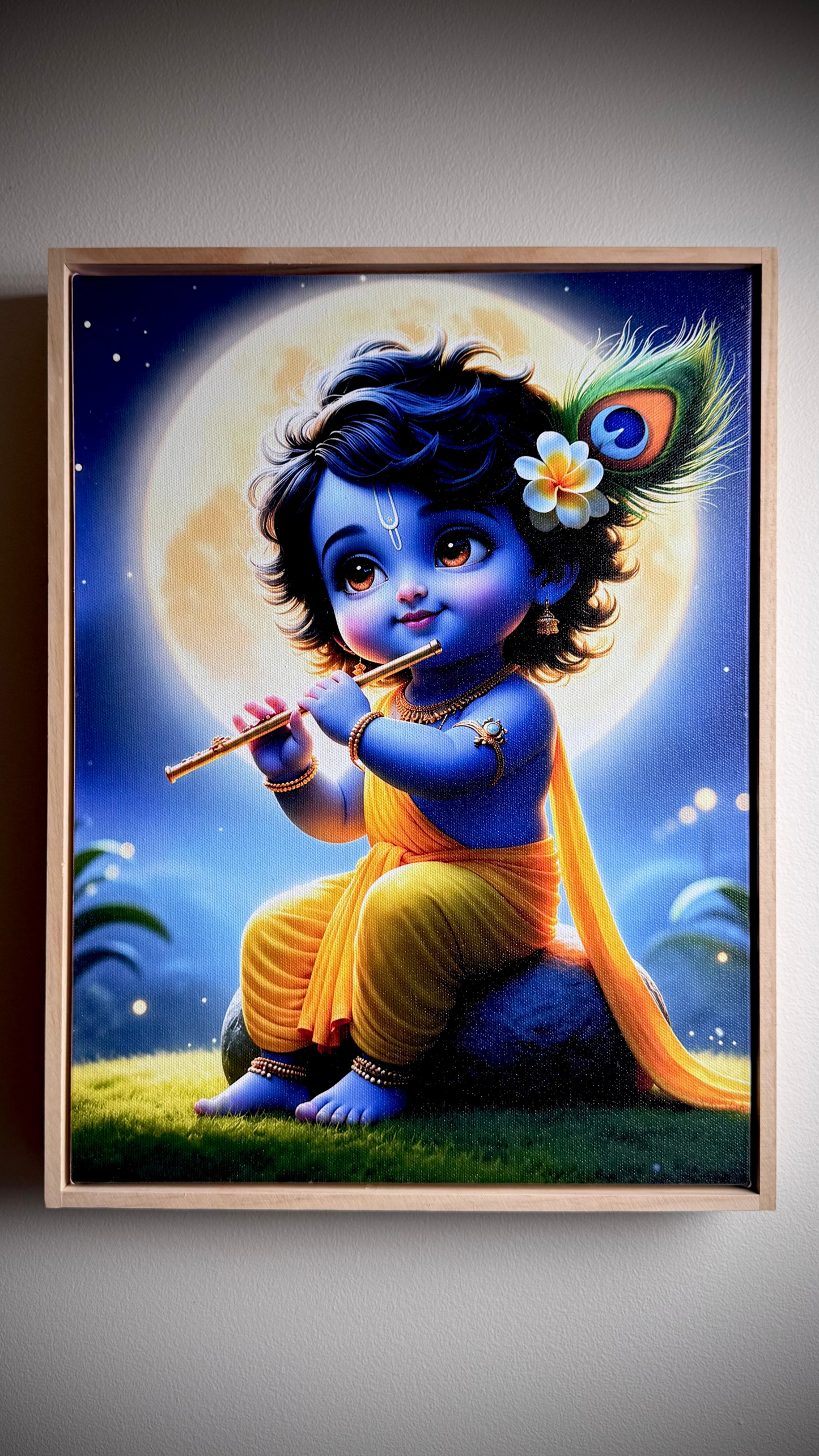 A framed art print depicting a stylized blue child playing a flute in front of a large moon, with a peacock feather flower and yellow drape, shown hanging on a gray wall.