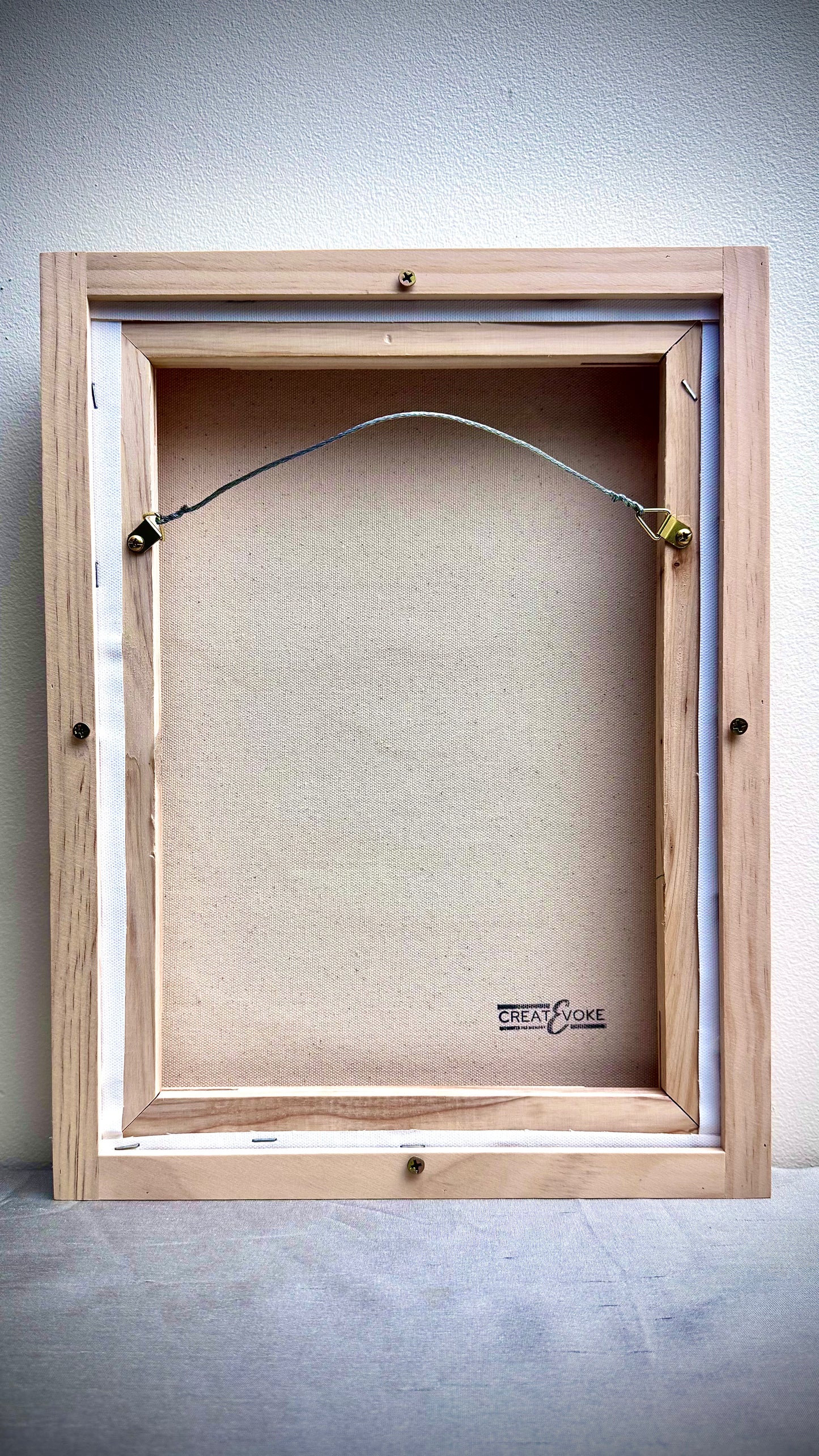 The back view of a wooden picture frame with a canvas mounted inside, showing the hanger wire and backing board against a neutral wall.